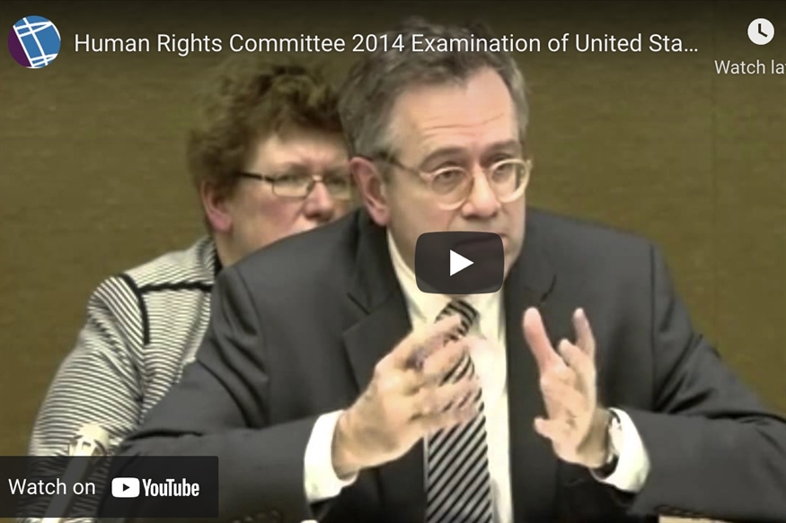 [ai] A man speaking during a session of the Human Rights Committee, with glasses and a black jacket, gesturing with his hands. A woman with curly hair in a striped shirt is visible in the background.